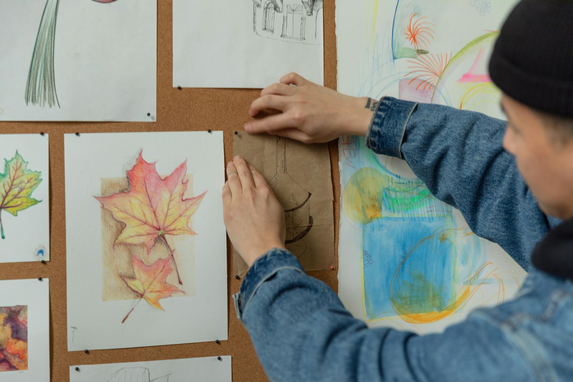 A person pins sketches on a corkboard, showcasing autumn leaf drawings in an artistic setup. -- pijushsaha.com