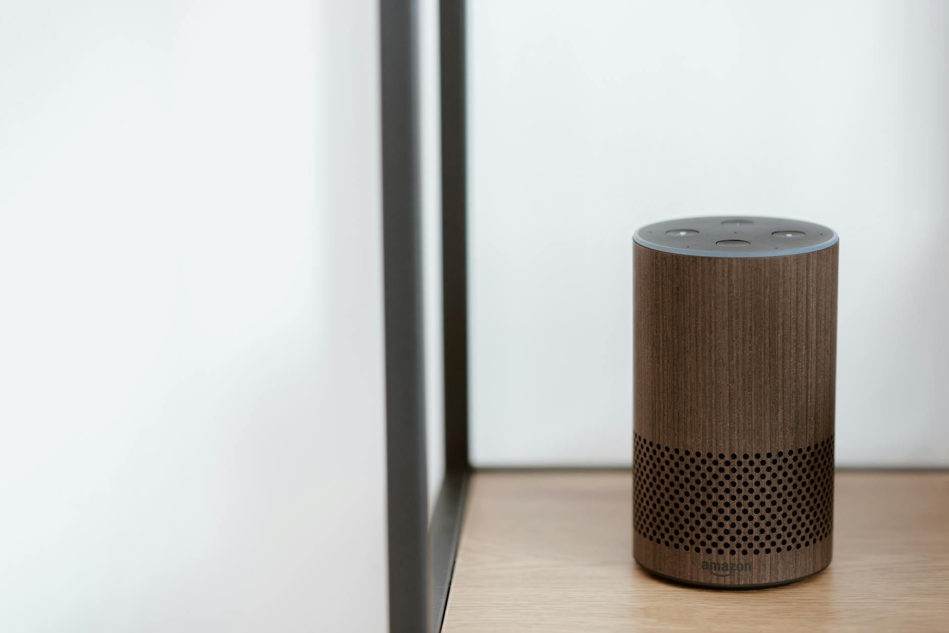 A sleek, modern smart speaker displayed on a wooden shelf in a minimalist room setting. — pijushsaha.com