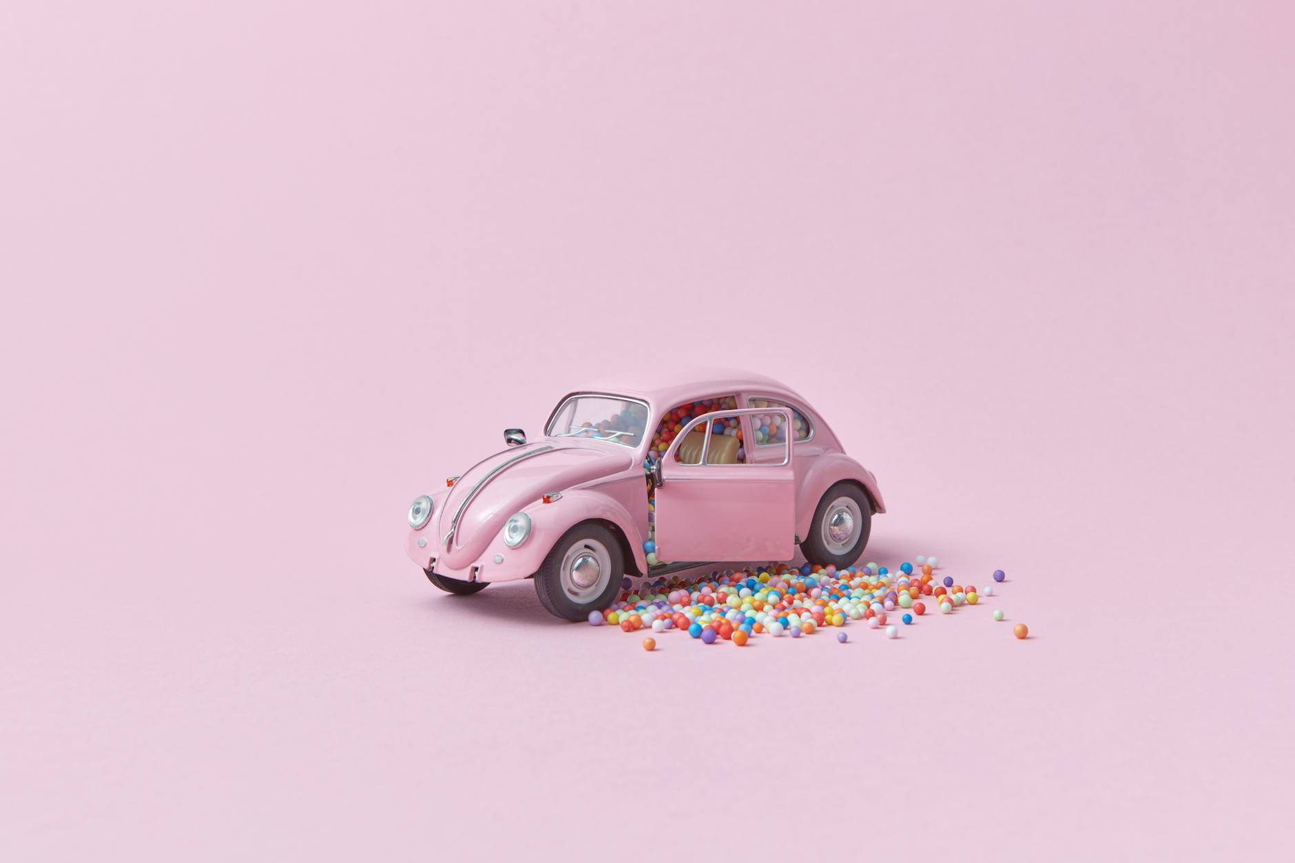 A pink toy car with open doors spilling colorful balls on a pastel pink background. -- pijushsaha.com