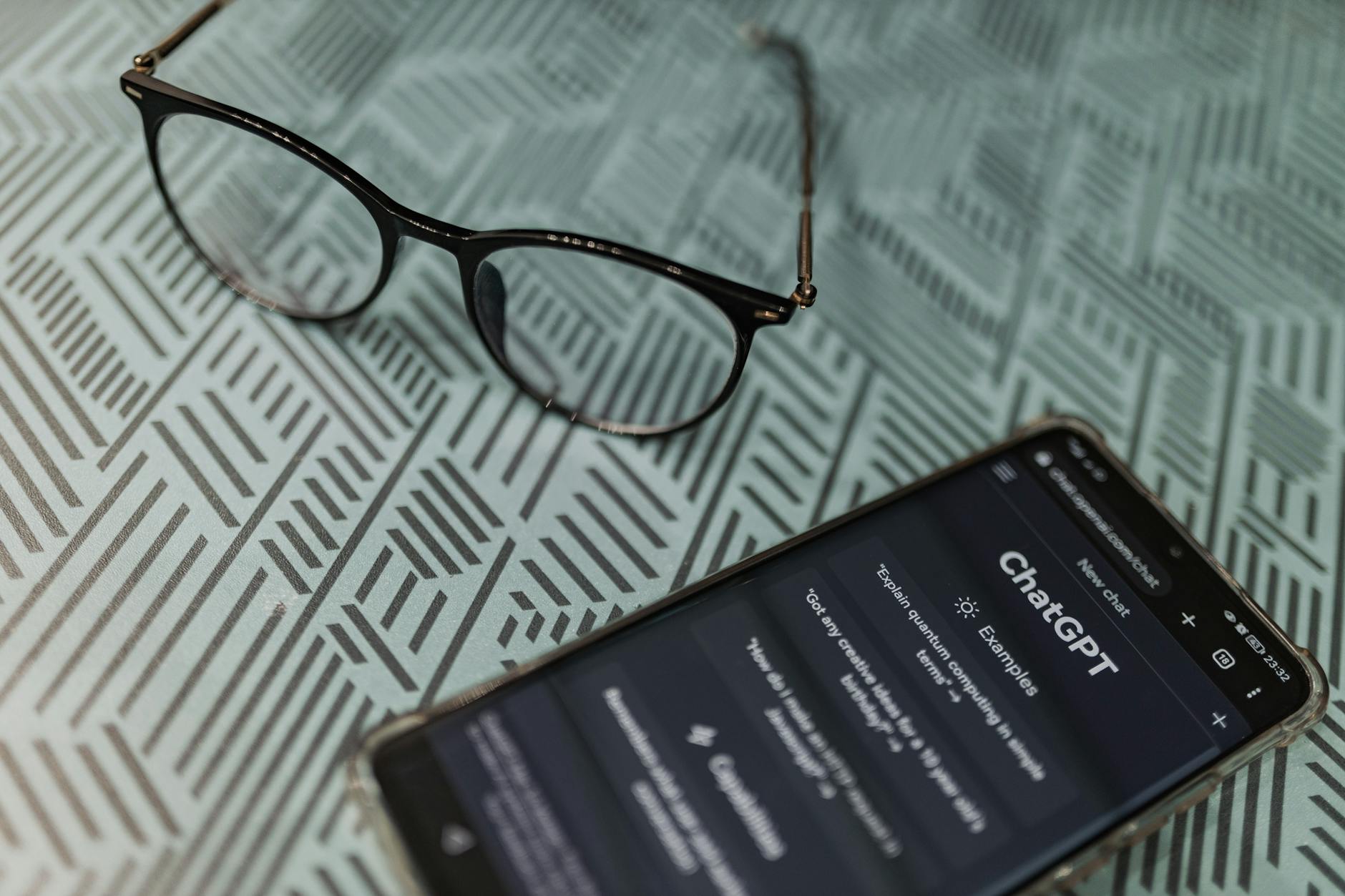 Eyeglasses next to a smartphone displaying the ChatGPT AI app on a patterned surface. — pijushsaha.com