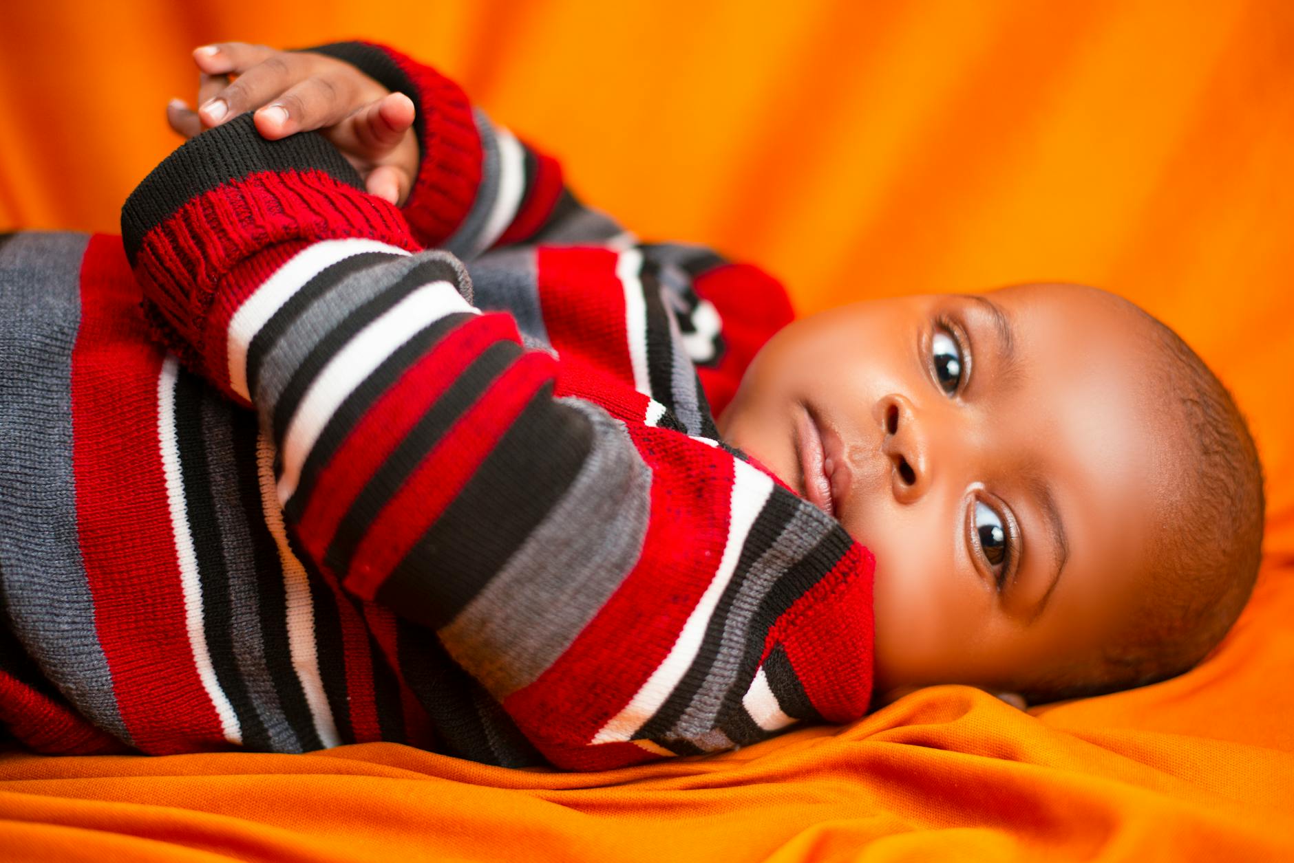 Adorable baby in striped sweater lying on an orange blanket, looking curious and content. -- pijushsaha.com